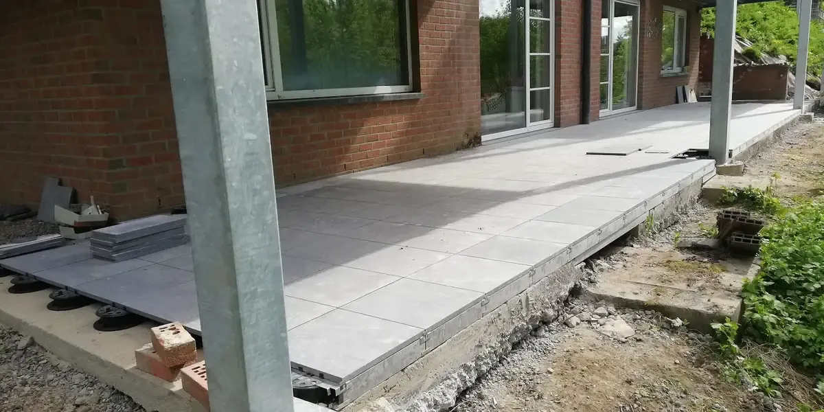 Terrasse en grandes dalles de carrelage gris posées sur plots le long d'une maison en briques, avec un poteau en acier galvanisé