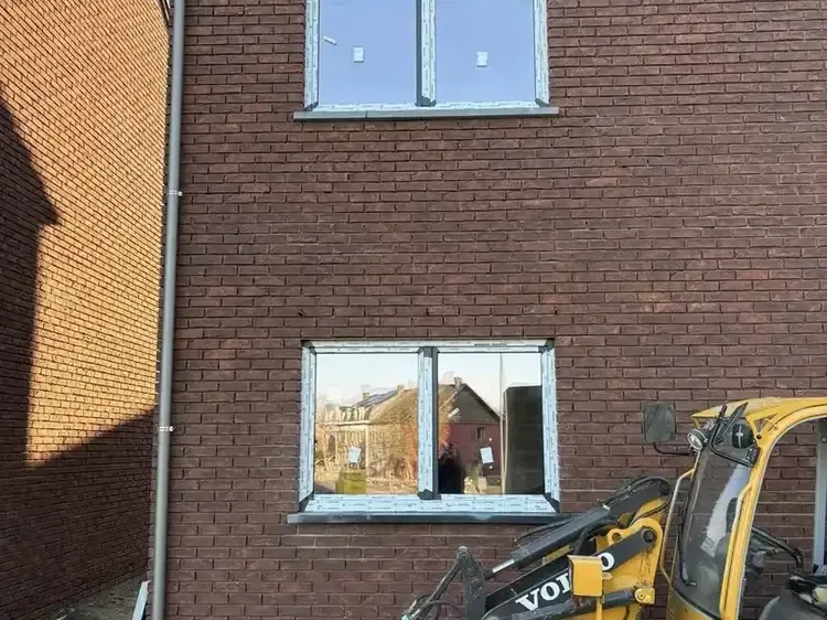 Façade en briques foncées avec deux châssis posés et un chargeur de chantier jaune au premier plan