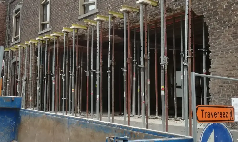 Façade en briques ouverte et soutenue par une rangée d'étais métalliques, benne bleue au premier plan