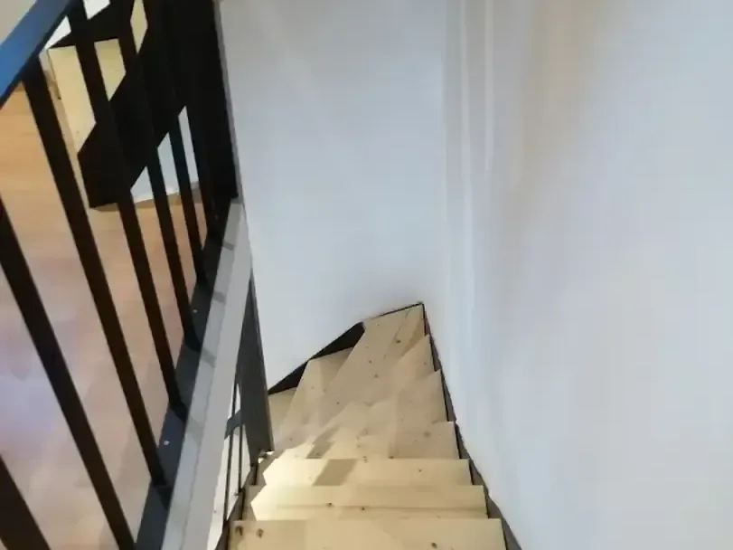 Vue plongeante sur un escalier en bois clair avec garde-corps en métal noir, dans une cage aux murs blancs