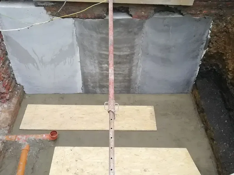 Fouille au pied d'un mur en briques avec parois en béton gris, sol coulé recouvert de panneaux de bois et tuyaux orange