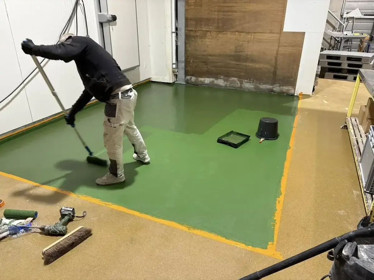 Ouvrier appliquant une résine verte au rouleau sur manche, bac de peinture et seau noir posés sur le sol