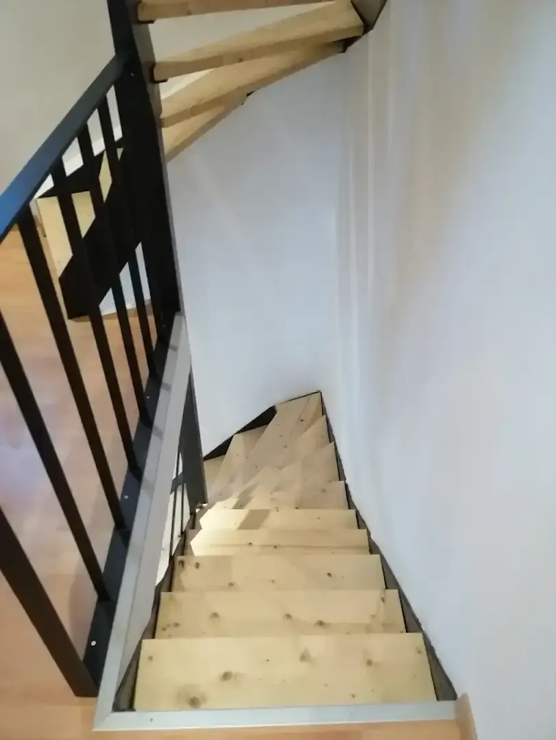 Vue plongeante sur un escalier en bois clair avec garde-corps en métal noir, dans une cage aux murs blancs
