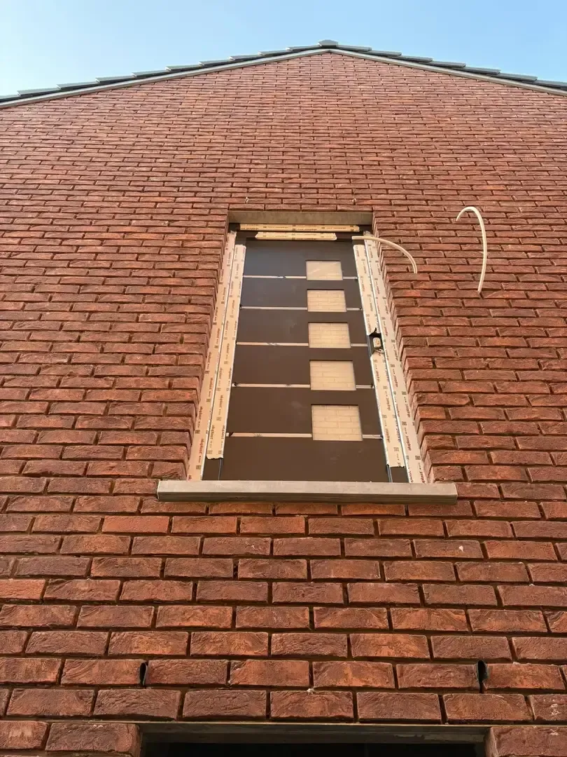 Façade en briques rouges avec une haute ouverture de fenêtre fermée par un panneau en bois, avant la pose du châssis