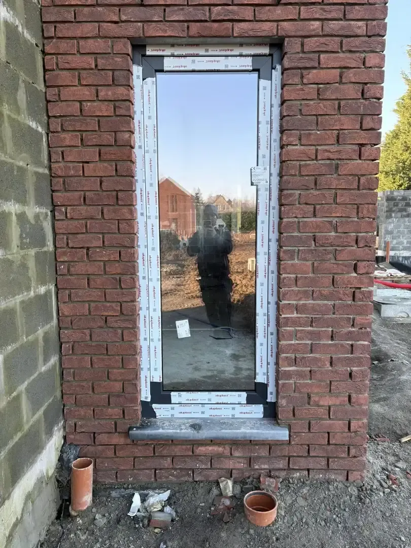 Ouverture étroite dans un mur de briques avec un châssis vitré posé, tuyaux orange au pied du mur