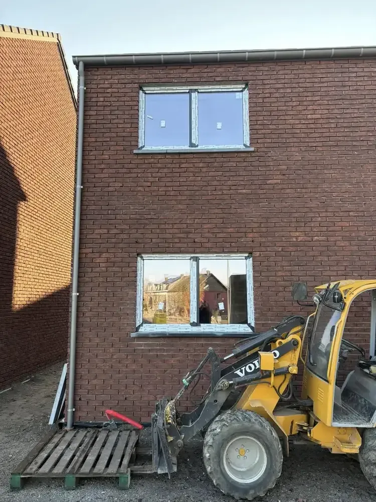 Façade en briques foncées avec deux châssis posés et un chargeur de chantier jaune au premier plan