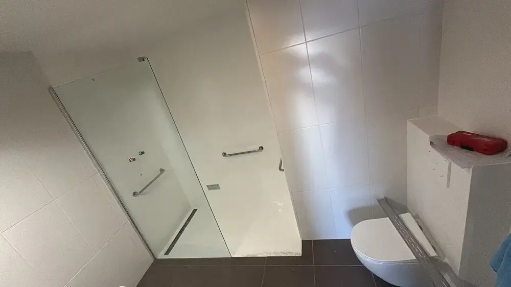 Salle d'eau avec douche vitrée, barres d'appui, caniveau au sol et WC suspendu