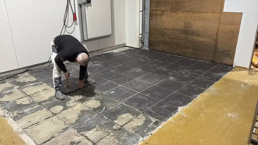 Ouvrier penché qui arrache d'anciens carreaux noirs, colle visible au sol et zone jaune déjà traitée à droite
