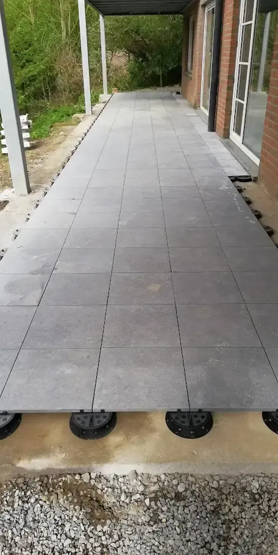 Allée en dalles de carrelage gris posées sur plots sous un auvent, le long d'une façade en briques à baies vitrées